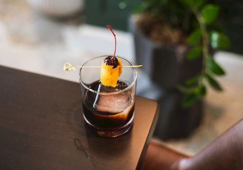 A neat cocktail on a dark table, garnished with an orange slice, a cherry, and a thin pick, with ice cubes in the glass.