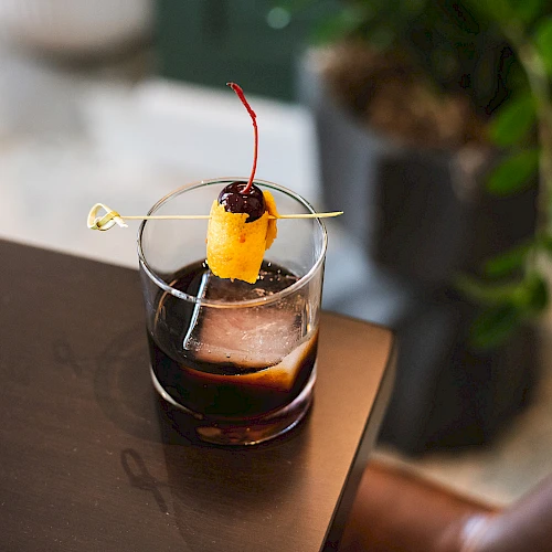 A neat cocktail on a dark table, garnished with an orange slice, a cherry, and a thin pick, with ice cubes in the glass.