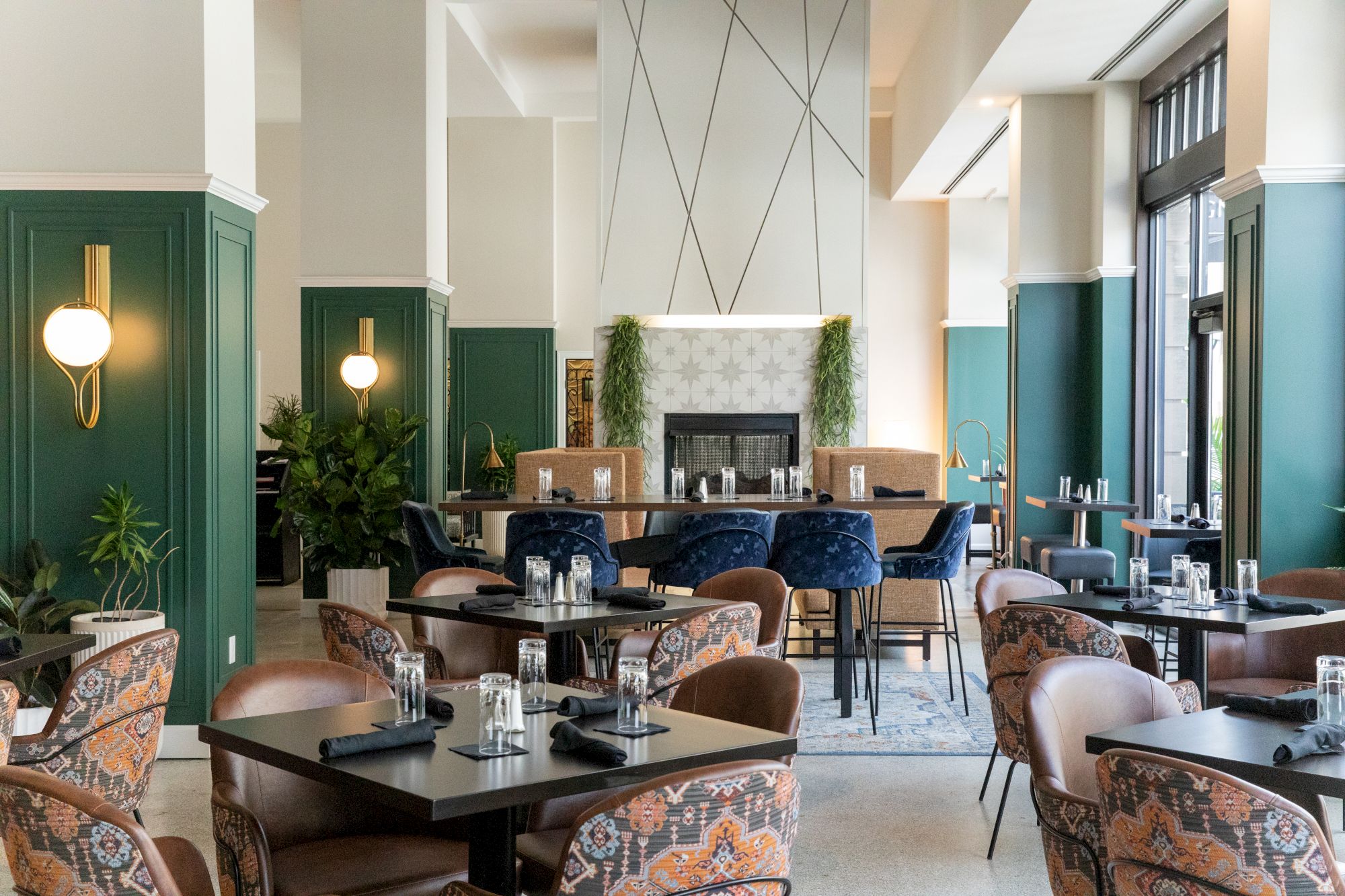 A stylish restaurant with green wall panels, modern decor, patterned chairs, set tables, and a central fireplace feature surrounded by plants.