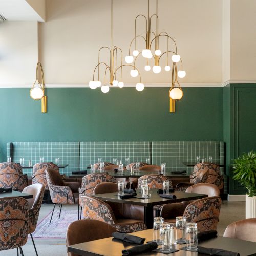 A modern restaurant with green walls, stylish chairs, and elegant light fixtures, featuring tables set with glassware and black napkins.