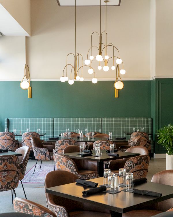 A modern restaurant with green walls, stylish chairs, and elegant light fixtures, featuring tables set with glassware and black napkins.