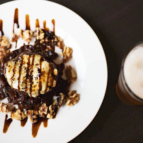 A plate with a gourmet dessert topped with grilled cheese, surrounded by walnuts and drizzled with chocolate sauce, next to a frothy glass of beer.