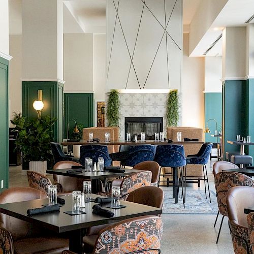 A modern, stylish restaurant interior with green walls, patterned chairs, and tables set for dining, featuring a fireplace in the background.