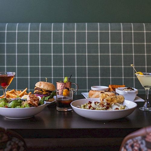 A table set with various dishes includes salads, a burger, fries, and a selection of cocktails, against a plaid-upholstered booth.