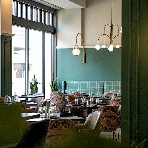 A stylish modern restaurant with green walls, large windows, elegant light fixtures, and patterned chairs. Tables are set, creating a cozy ambiance.