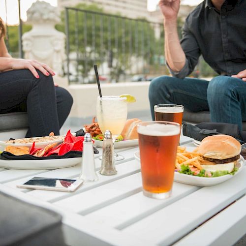 Two people are sitting at an outdoor table with burgers, fries, drinks, and sunglasses. A phone and a salt shaker are also on the table.