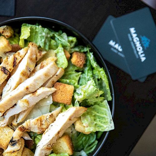 A bowl of salad topped with grilled chicken strips and croutons is next to two black cards with "Magnolia" written on them.