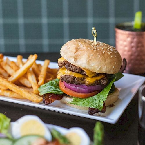 A plate with a double cheeseburger, lettuce, tomato, red onion, bacon, and fries is on a table with drinks.