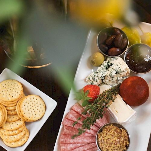 The image shows a charcuterie board with cheese, meats, olives, mustard, pickles, crackers, and a strawberry, displayed on white dishes.