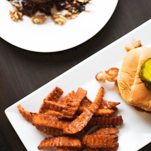 The image shows a meal consisting of a burger with a pickle on top, crinkle-cut fries, another dish in the background, and a glass of beer.