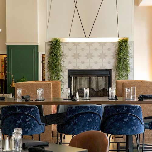 A modern dining area with blue velvet chairs, a long wooden table set with glasses, and a decorative fireplace adorned with plants and tile pattern.