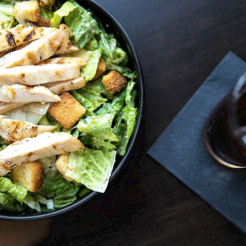 A bowl of Caesar salad with grilled chicken and croutons, alongside a glass of iced drink with a lemon slice on a dark napkin.