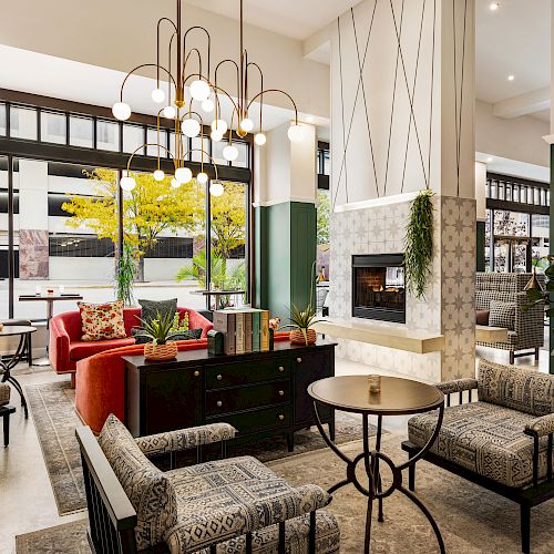 A modern living room with stylish furniture, plants, large windows, and a fireplace against a tiled wall, illuminated by elegant lighting fixtures.