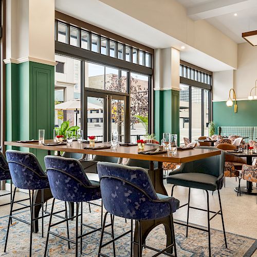 An elegant modern dining area with high ceilings, pendant lights, blue velvet barstools, and large windows providing a view of the outdoors.