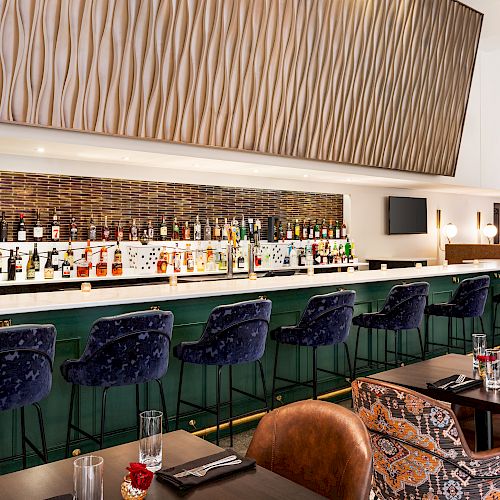 A modern bar with a variety of spirits, blue bar stools, cushioned seating, decorative wall paneling, and a sleek green and white interior.