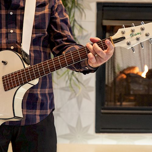 A person is playing an electric guitar in front of a lit fireplace, wearing a plaid shirt. The scene appears cozy and relaxing.