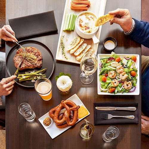 A table with two people enjoying various dishes including steak, asparagus, salad, bread, pretzels with mustard and cheese, and drinks like wine, and a beer.