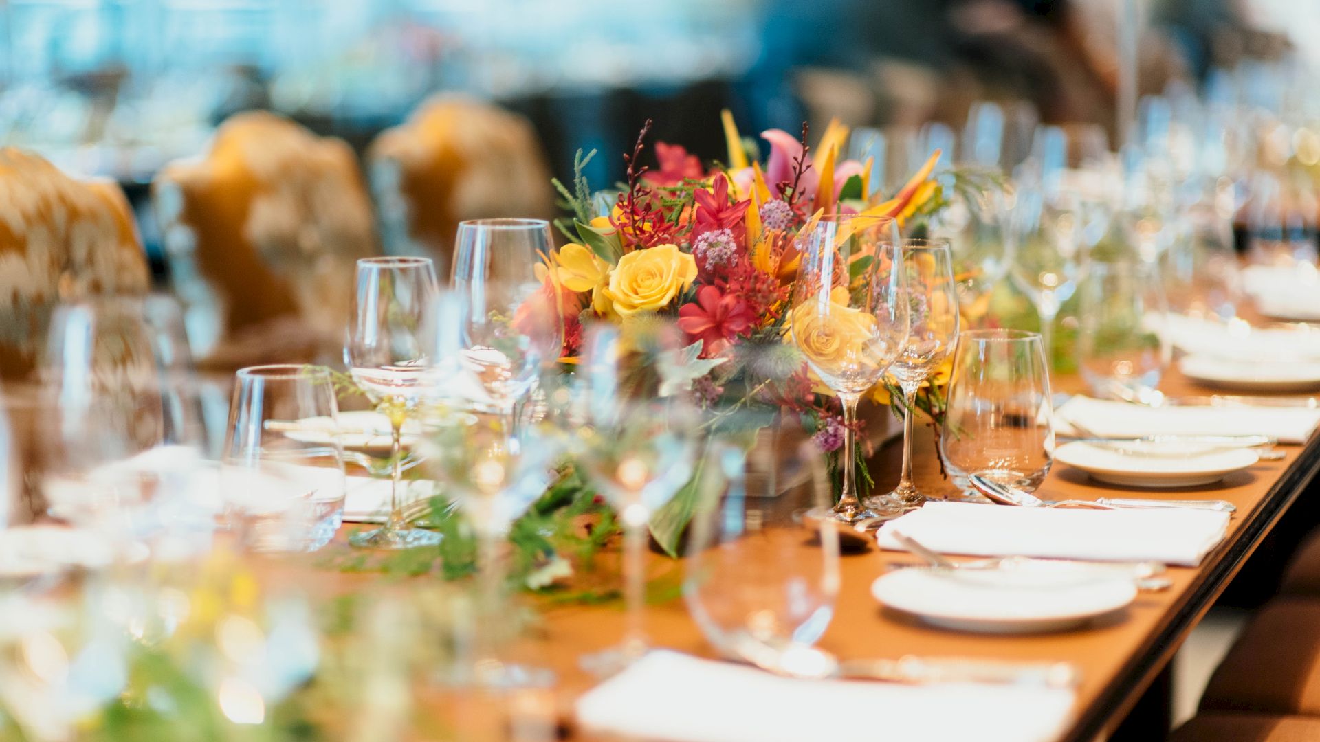 A long dining table is elegantly set with glassware, napkins, and bright floral centerpieces, suggesting a formal event or celebration.