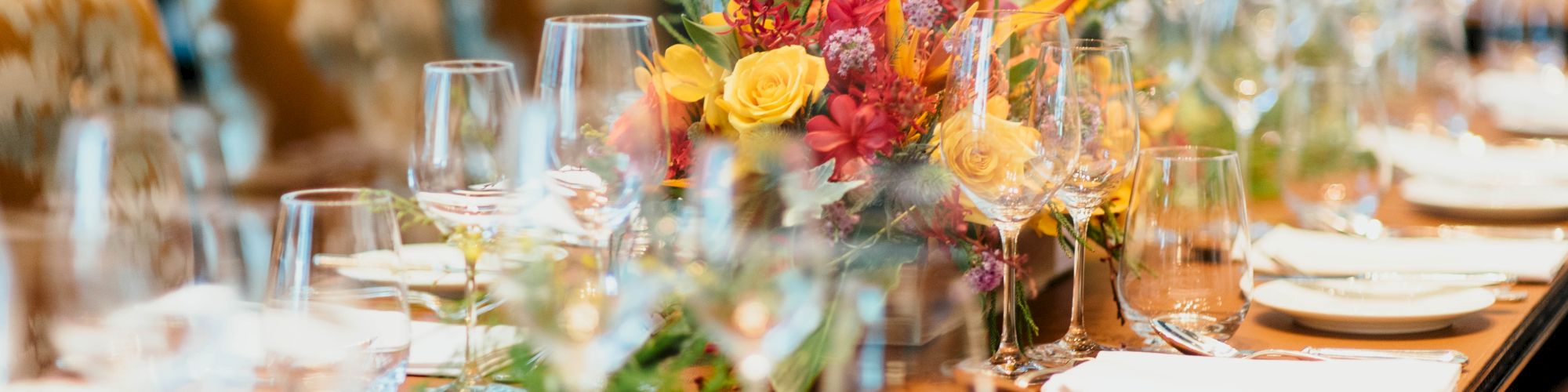 A long dining table is elegantly set with glassware, napkins, and bright floral centerpieces, suggesting a formal event or celebration.