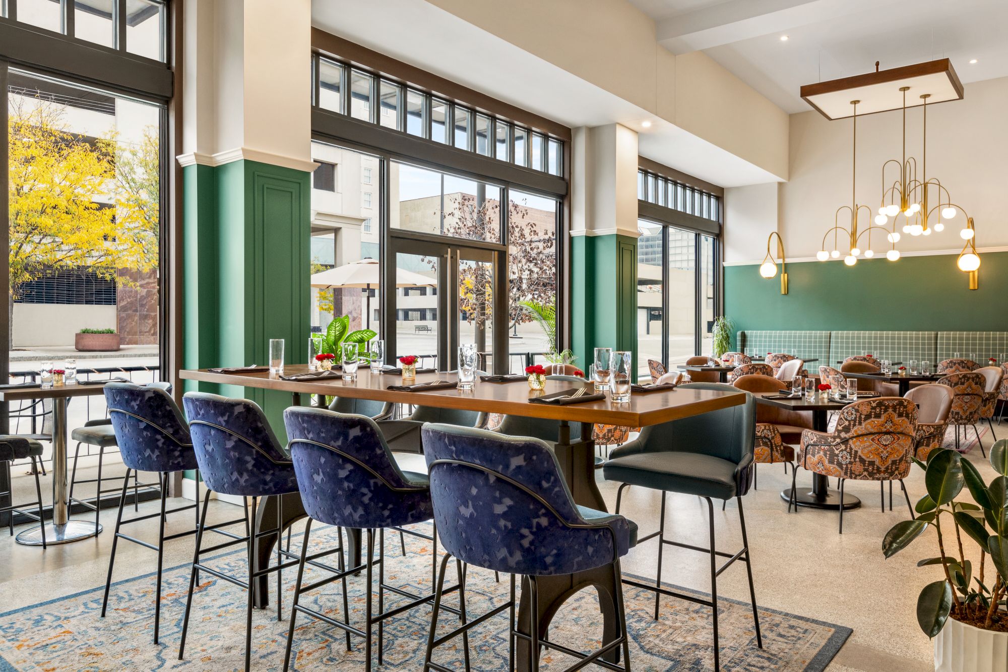 The image shows a stylish, modern restaurant with high ceilings, blue upholstered chairs, large windows, and elegant lighting fixtures.