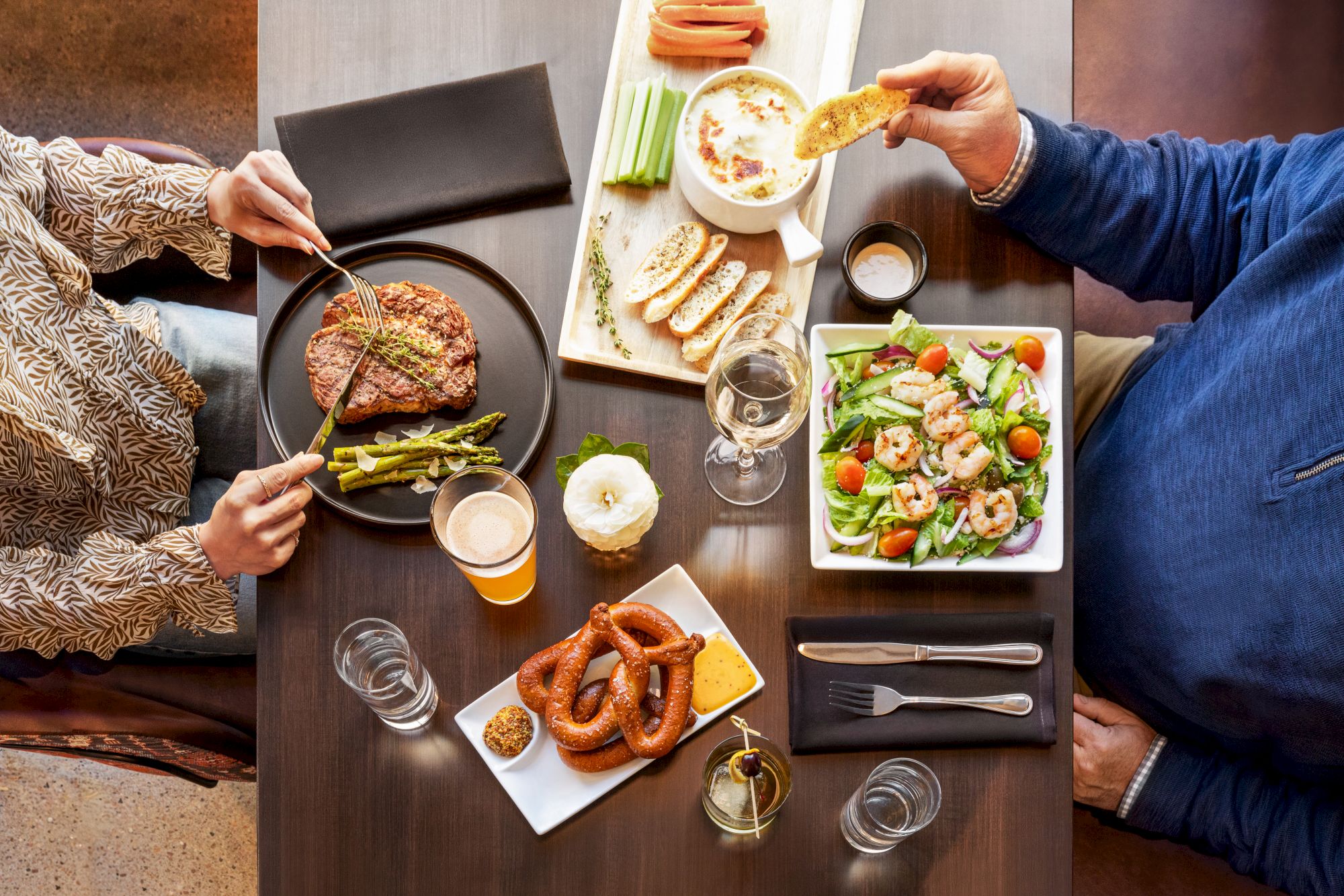 Two people dining with a table set with a steak dish, salad, pretzels, dipping sauce, vegetable platter, bread, wine, and a candle centerpiece.