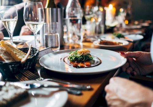 A dinner table is set with plates of gourmet food, wine glasses, and bread, with people seated and enjoying the meal in a dimly lit ambiance.