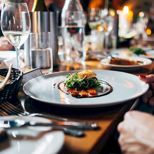 A dinner table is set with plates of gourmet food, wine glasses, and bread, with people seated and enjoying the meal in a dimly lit ambiance.