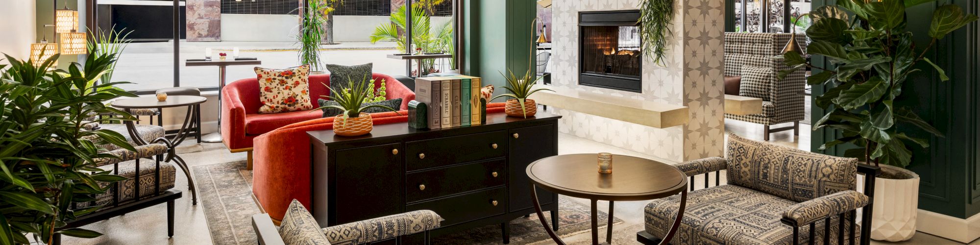 A stylish seating area with modern furniture, a fireplace, and plants. It features large windows allowing natural light to enter the space.