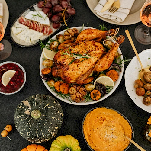 The image shows a Thanksgiving meal with a roast turkey, mushrooms, cranberries, cheese, grapes, and wine, garnished with flowers and pumpkins.