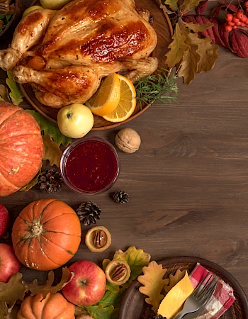 A Thanksgiving-themed spread with roasted chicken, pumpkins, apples, oranges, nuts, cranberries, and autumn leaves on a wooden table.