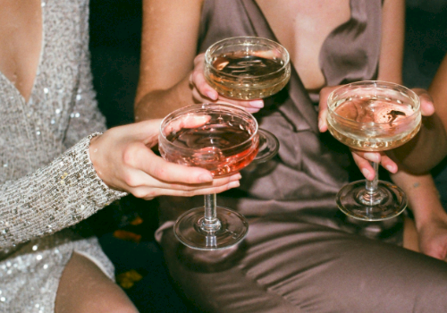 Three people in elegant outfits holding champagne glasses with drinks, appearing to be in a celebratory or festive setting.