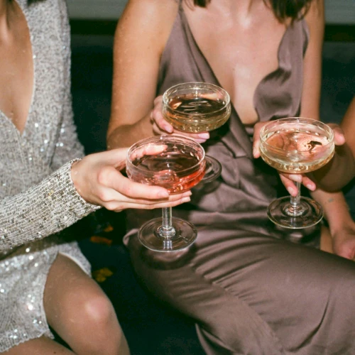 Three people in elegant outfits holding champagne glasses with drinks, appearing to be in a celebratory or festive setting.