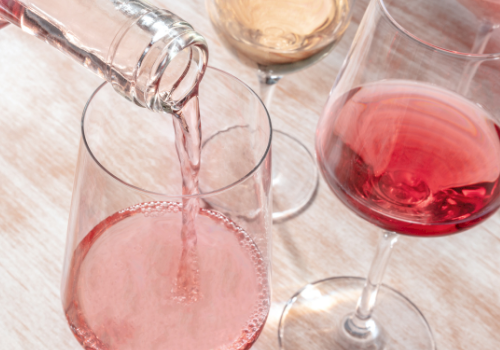 A bottle is pouring rosé wine into one of several wine glasses on a wooden surface.
