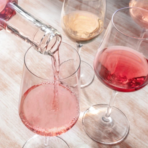 A bottle is pouring ros&eacute; wine into one of several wine glasses on a wooden surface.