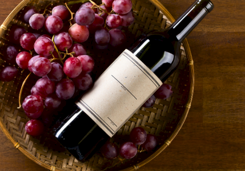 A wine bottle and red grapes sit in a woven basket on a wooden surface.