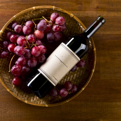 A wine bottle and red grapes sit in a woven basket on a wooden surface.