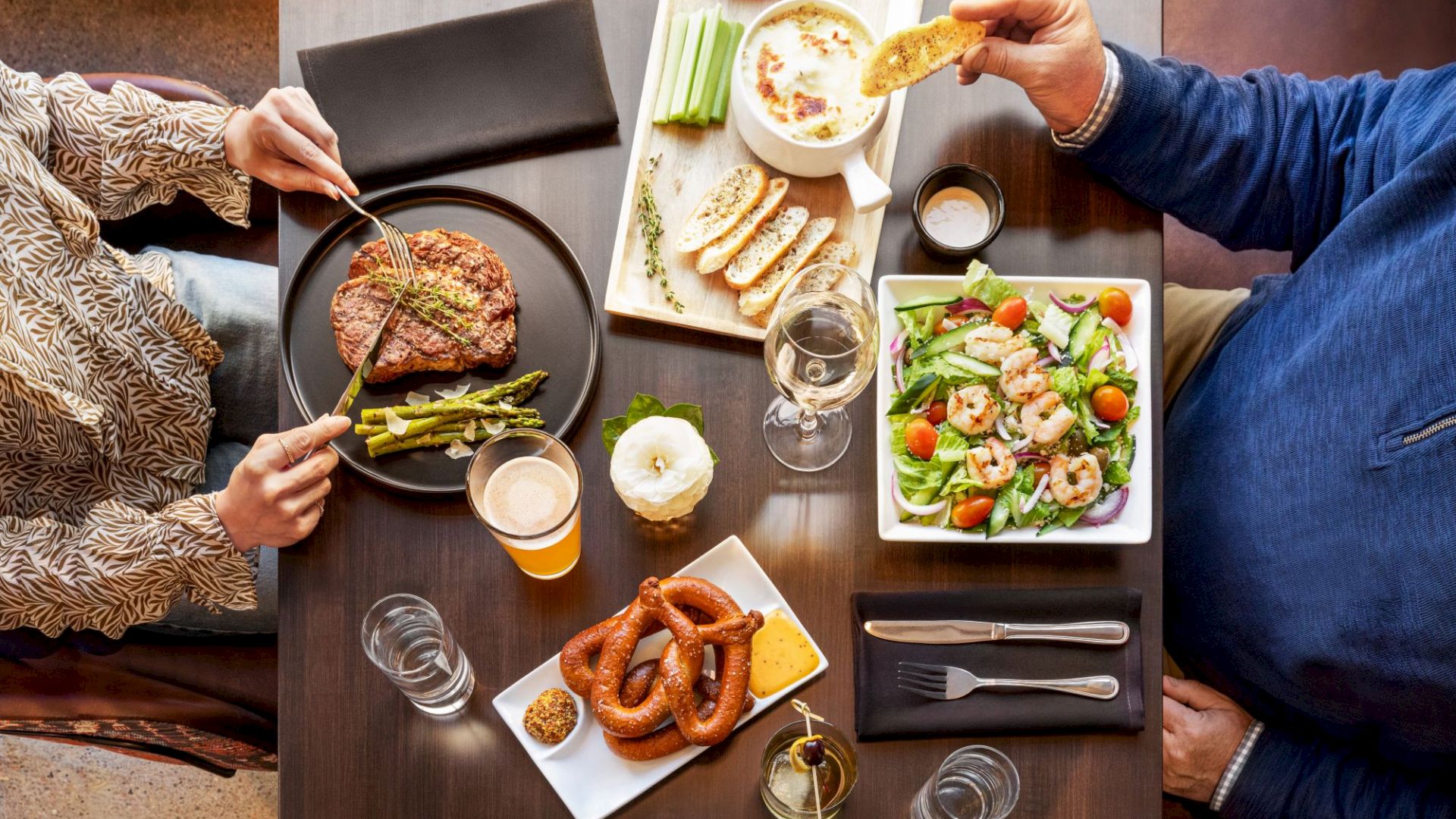 Friends share a hearty spread: steak, asparagus, salad, bread and sausages, with wine and beer on the table.
