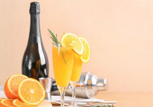 A bottle of sparkling wine, two fluted glasses with orange slices, fresh orange halves, a rosemary sprig, and a citrusy cocktail setup on a tray.