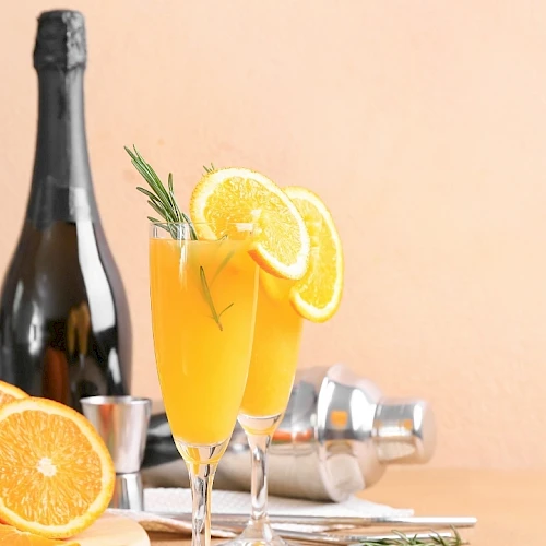 A bottle of sparkling wine, two fluted glasses with orange slices, fresh orange halves, a rosemary sprig, and a citrusy cocktail setup on a tray.